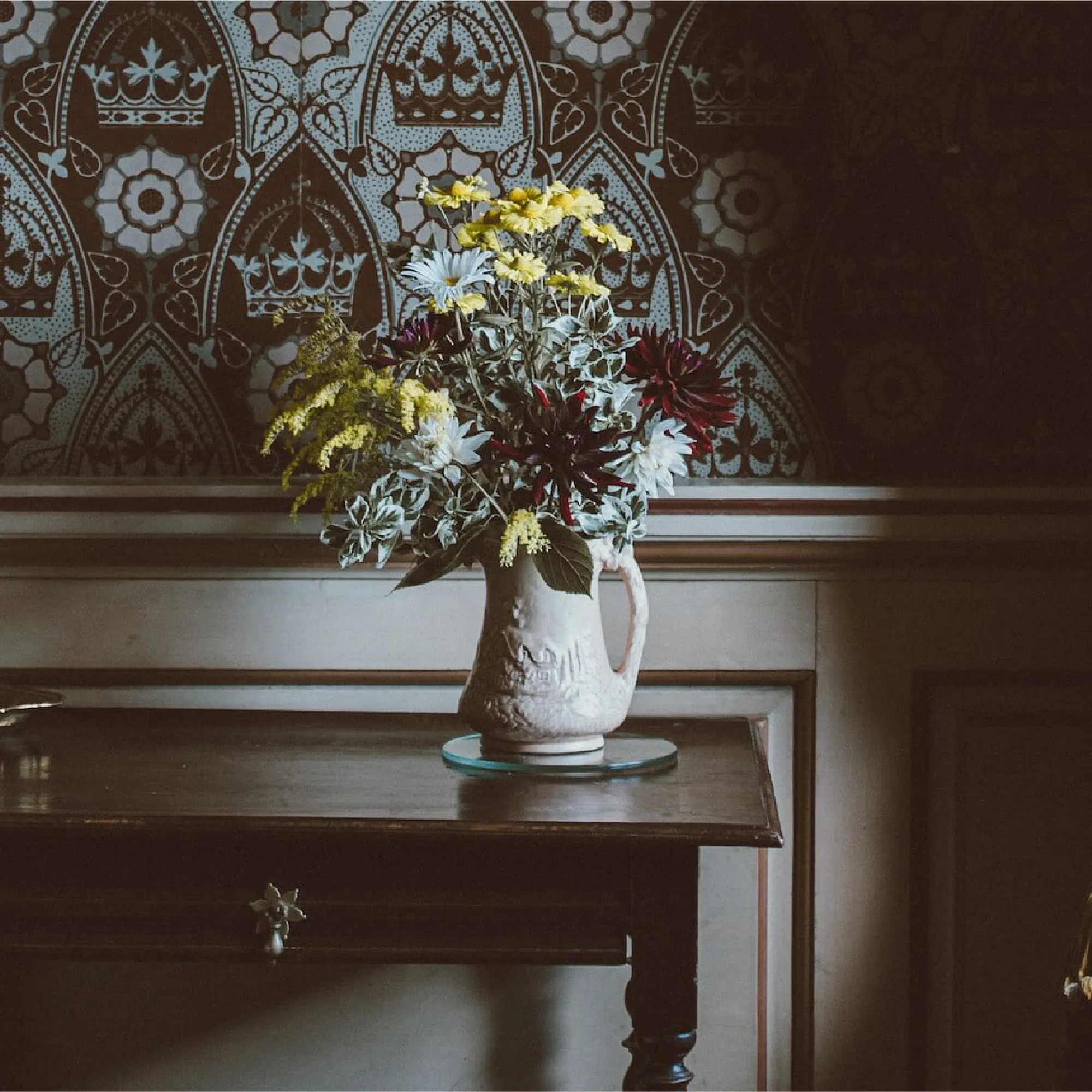A vase of flowers and a candlestick on a dark wooden table in front of a medieval-looking interior with wallpaper and a heavy curtain tied back with a rope with tassels on the end.