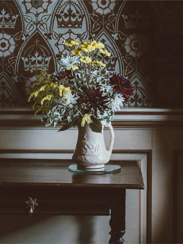 A vase of flowers and a candlestick on a dark wooden table in front of a medieval-looking interior with wallpaper and a heavy curtain tied back with a rope with tassels on the end.